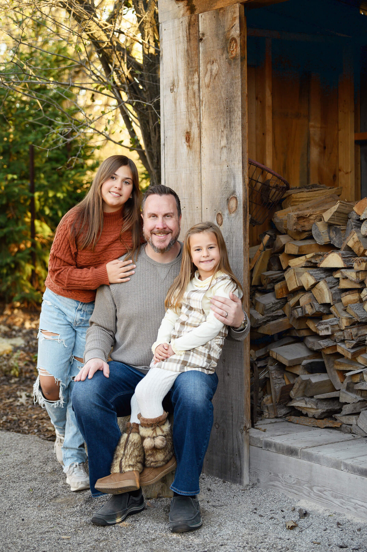 dad with his two daughters sitting beside a wood shed for their fall family photos in Burlington, ON