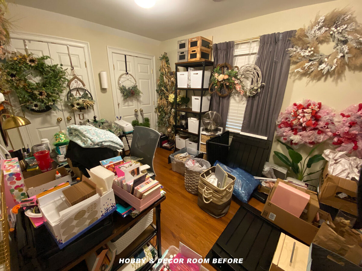 A cluttered hobby and decor room filled with various items and craft materials. Wreaths hang on the walls and doors, and boxes and containers are scattered across the floor and desk. A large flower decoration is beside the window with gray curtains.