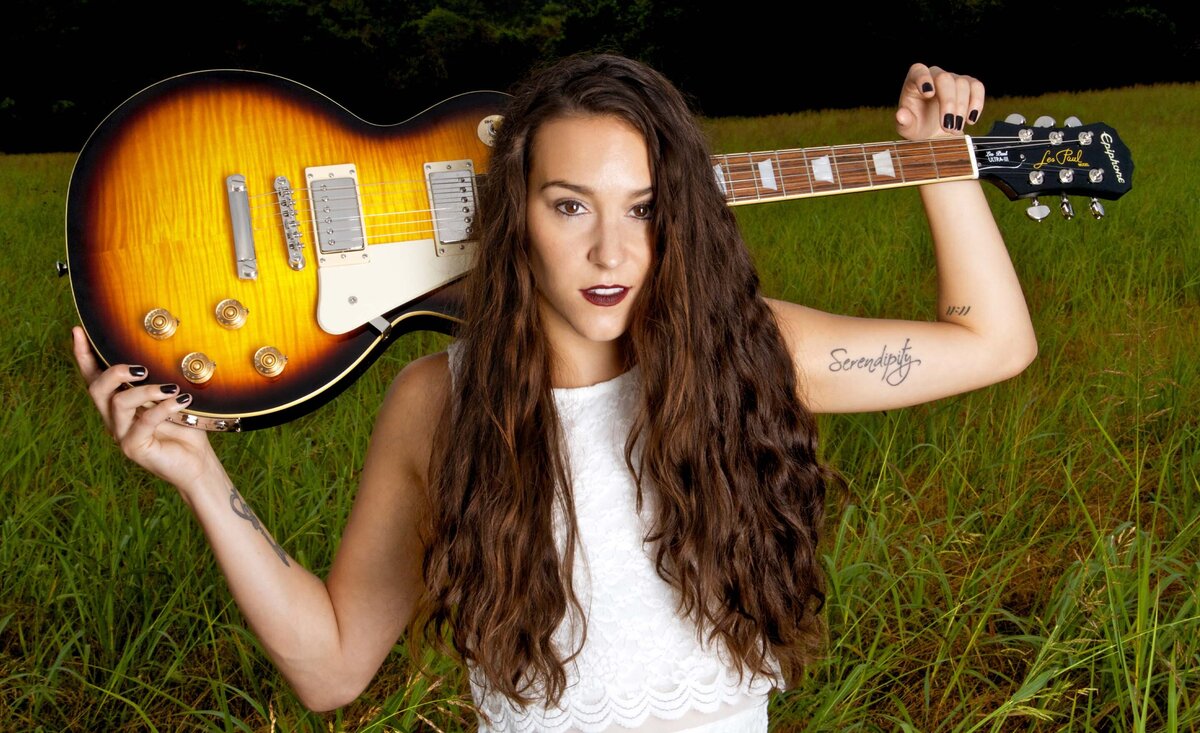 Female musician portrait Leanne Pearson wearing white dress holding sunburst electric guitar behind head while standing against tall green grass background