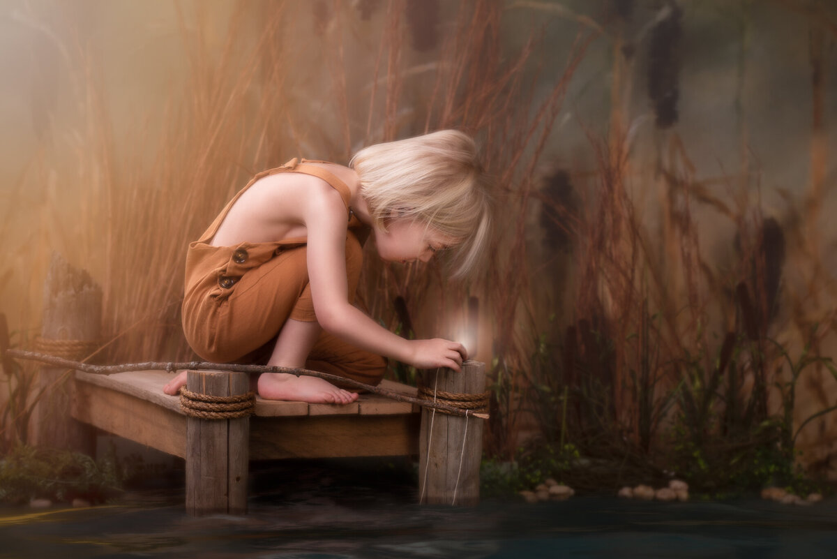 Fine art portrait of a young child sitting on a wooden dock surrounded by natural reeds, deeply immersed in a moment of exploration. Captured in Overland Park, Kansas, this serene and nature-inspired setup is perfect for families seeking creative and rustic child photography.