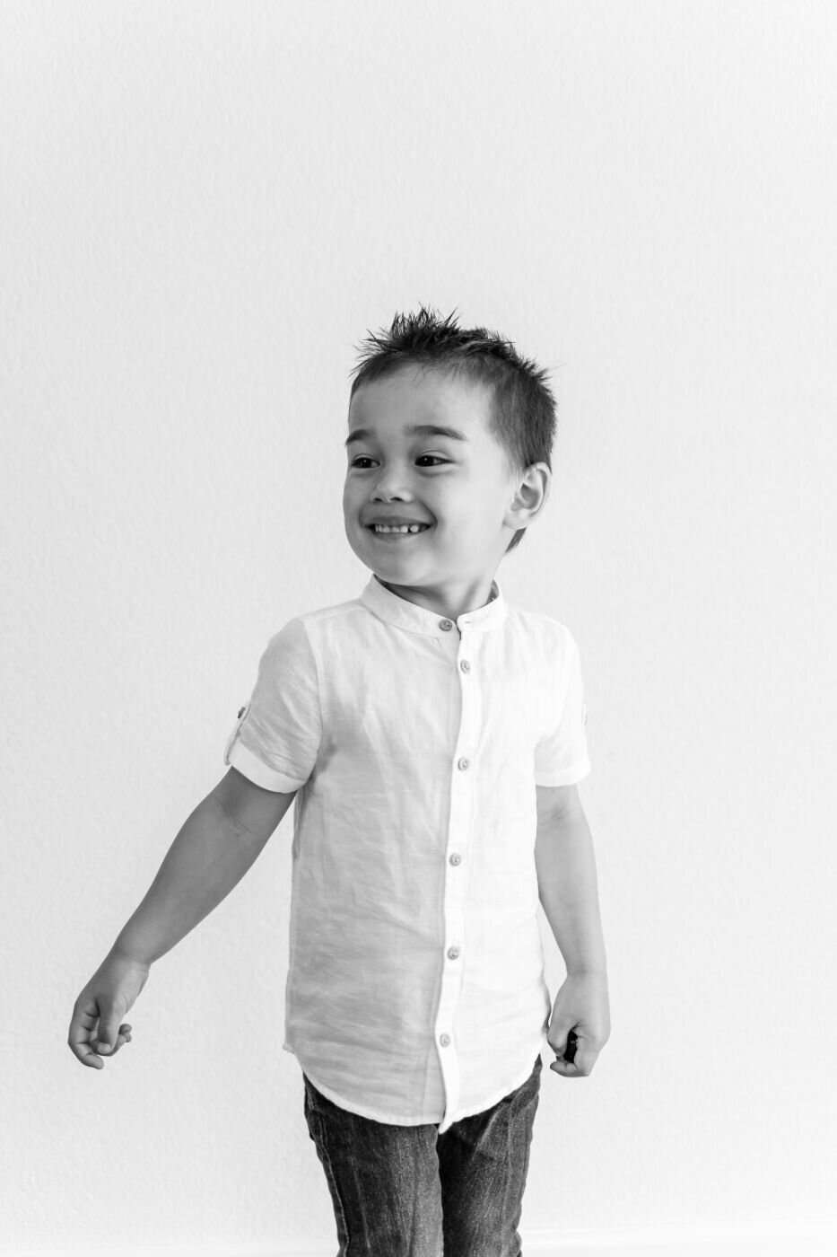A candid, black and white portrait of a young boy smiling and looking to the side. He is wearing a short-sleeved white button-down shirt and dark pants. His dark hair is styled in a spiky faux hawk. He stands against a simple white background.
