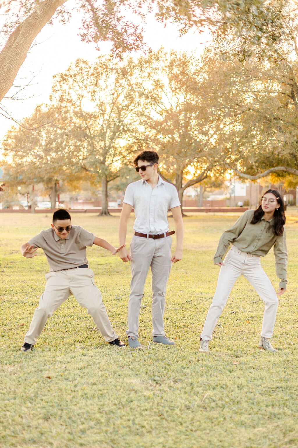 three young people posing playfully outdoors in a grassy park at sunset. The person in the center stands tall, striking a confident, flexing pose while wearing sunglasses, a light short-sleeve button-up shirt, light gray pants, and blue shoes. On the left, a boy in a gray polo shirt and beige pants strikes a dynamic, crouched action pose, pretending to punch the center person’s arm. On the right, a girl in a green button-up shirt and white pants leans toward the center, smiling and mirroring the playful energy. Trees with golden sunlight filtering through their leaves create a warm and cheerful backdrop