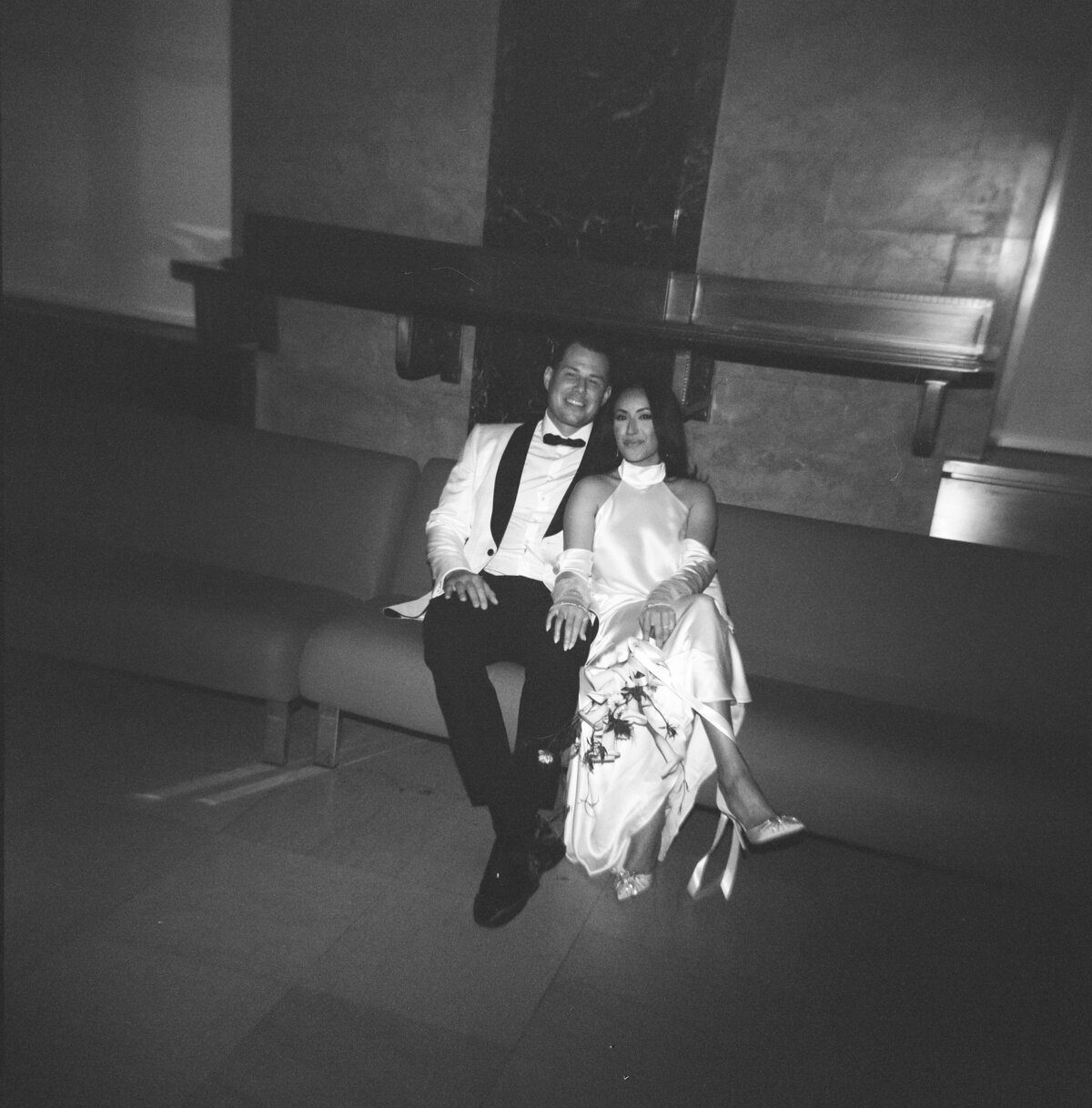 Black and white film photo of the bride and groom sitting together on a bench inside New York City Hall, captured during Japna and Chris’s intimate elopement by NYC film wedding photographer Perry Hancock.