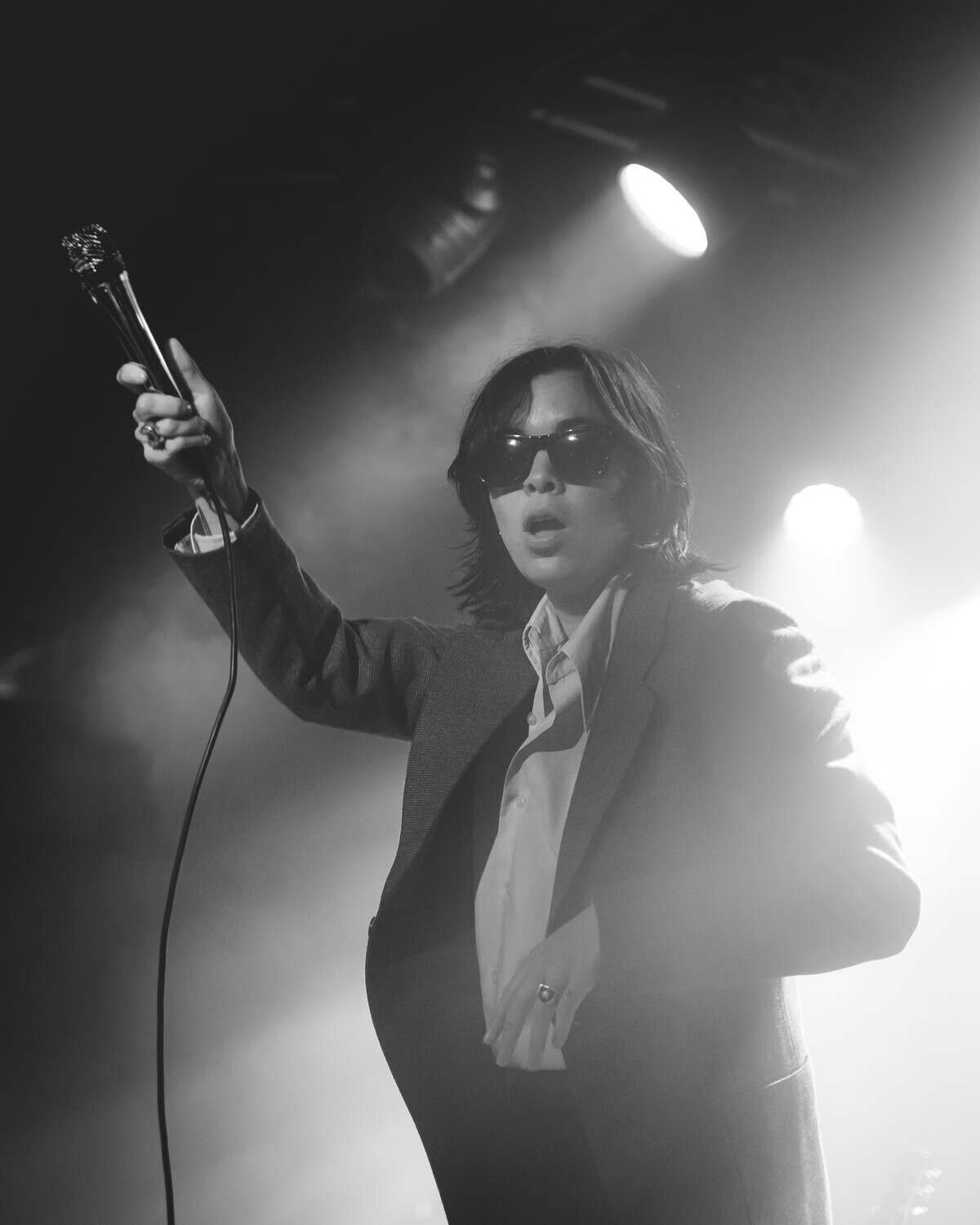 The aces singer holds mic aloft on stage and poses with sunglasses on