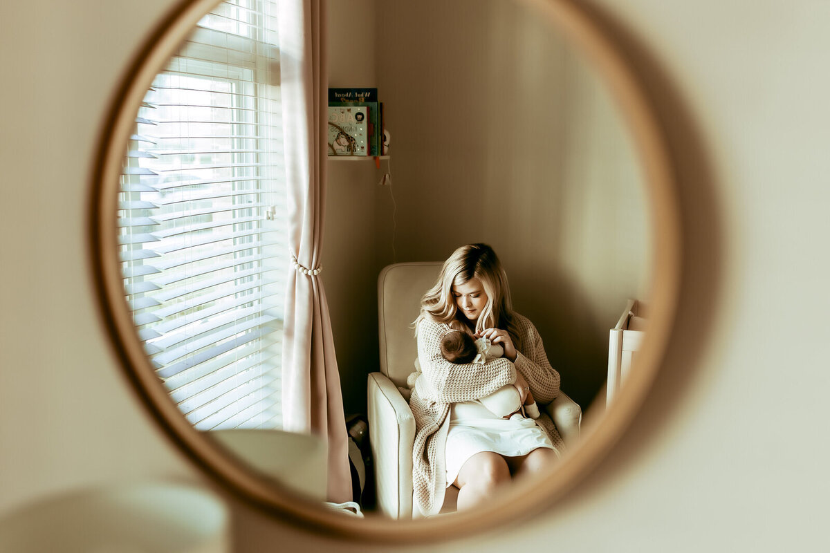 A woman sitting in a chair breastfeeding a baby is reflected in a round mirror on the wall. The room has a window with blinds and a small shelf with books.