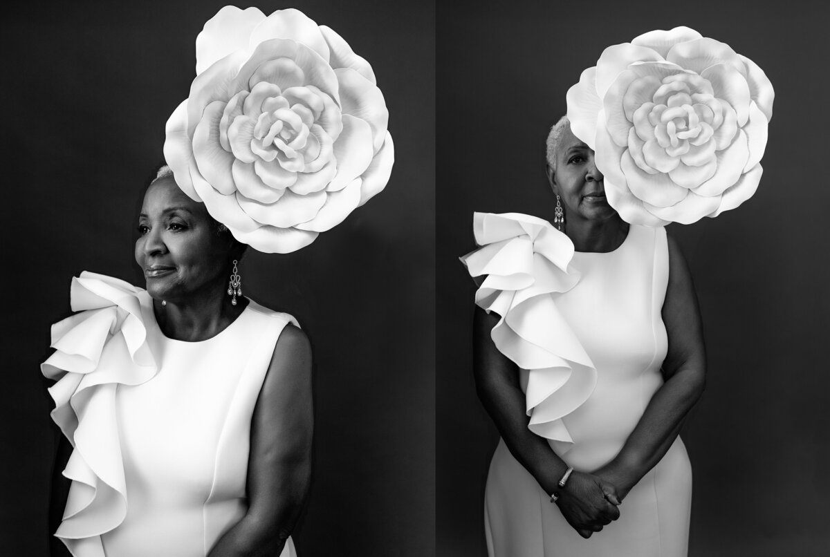 A sophisticated portrait from a Cincinnati studio, showcasing a woman in her late 60s wearing a stylish white gown with a ruffled shoulder, poised against a black backdrop with a striking white flower headpiece, radiating elegance and strength.
