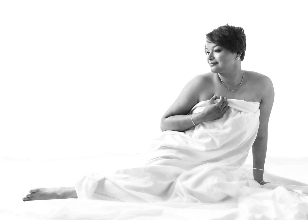 black and white boudoir woman of woman sitting and covered in white bedsheet