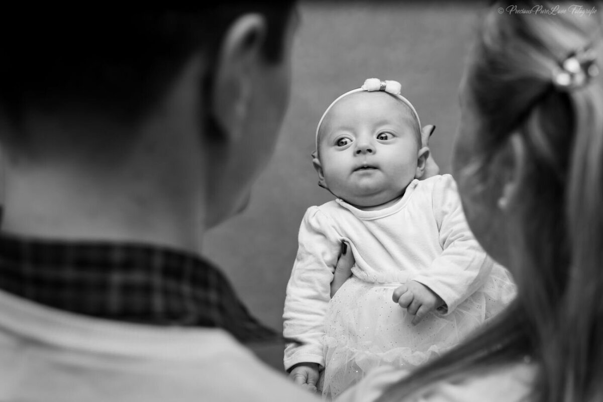  A black and white, eye-level photo of a newborn baby being held up between two parents. The parents are out of focus in the foreground, looking at the baby. The baby is in sharp focus, looking directly at the camera with wide eyes. The baby is wearing a white outfit and a small bow on its head.