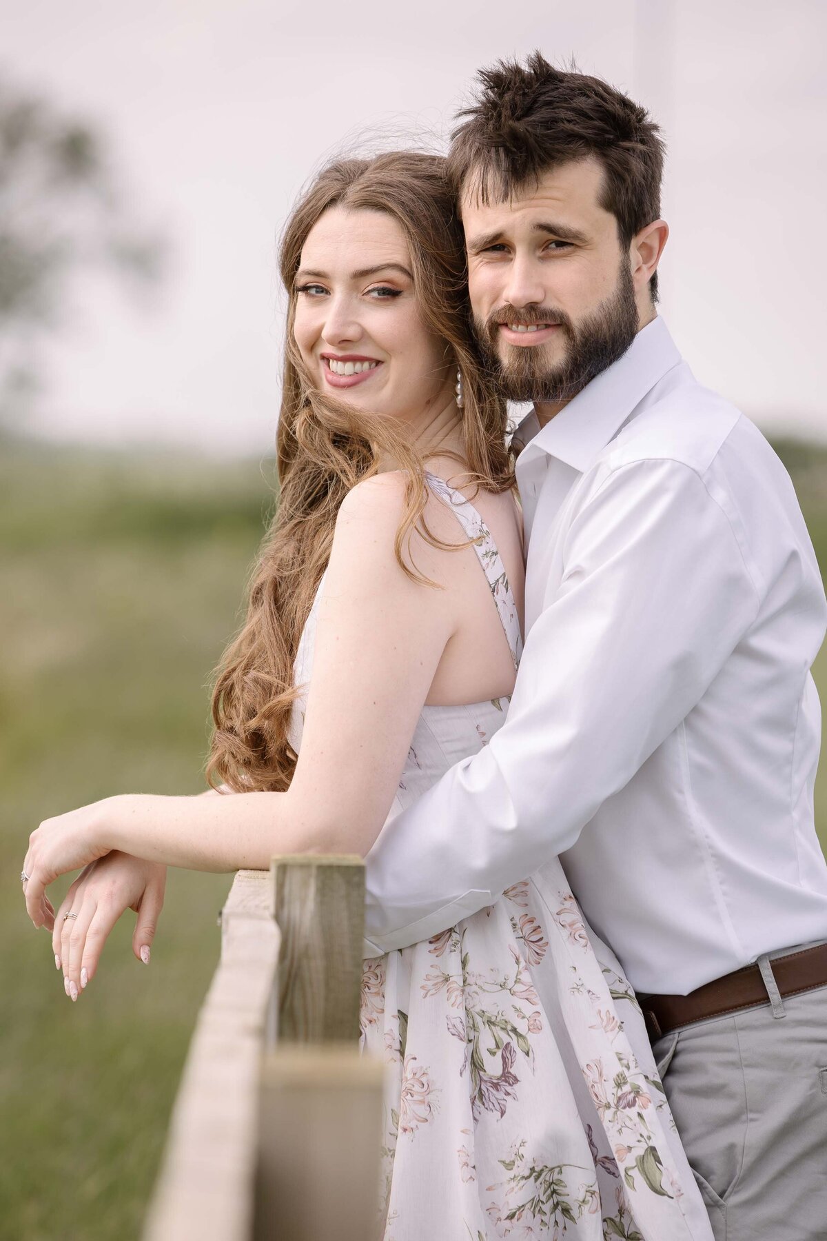 An engaged couple resting on a fence looking at the camera