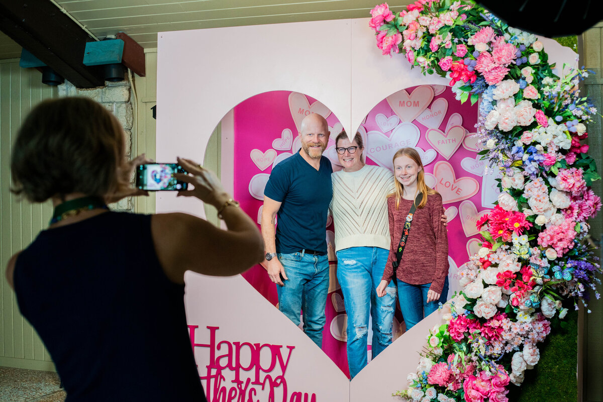 Woman taking a photo of a family posing inside of a heart shaped photo booth Mother's Day backdrop decorated with flowers