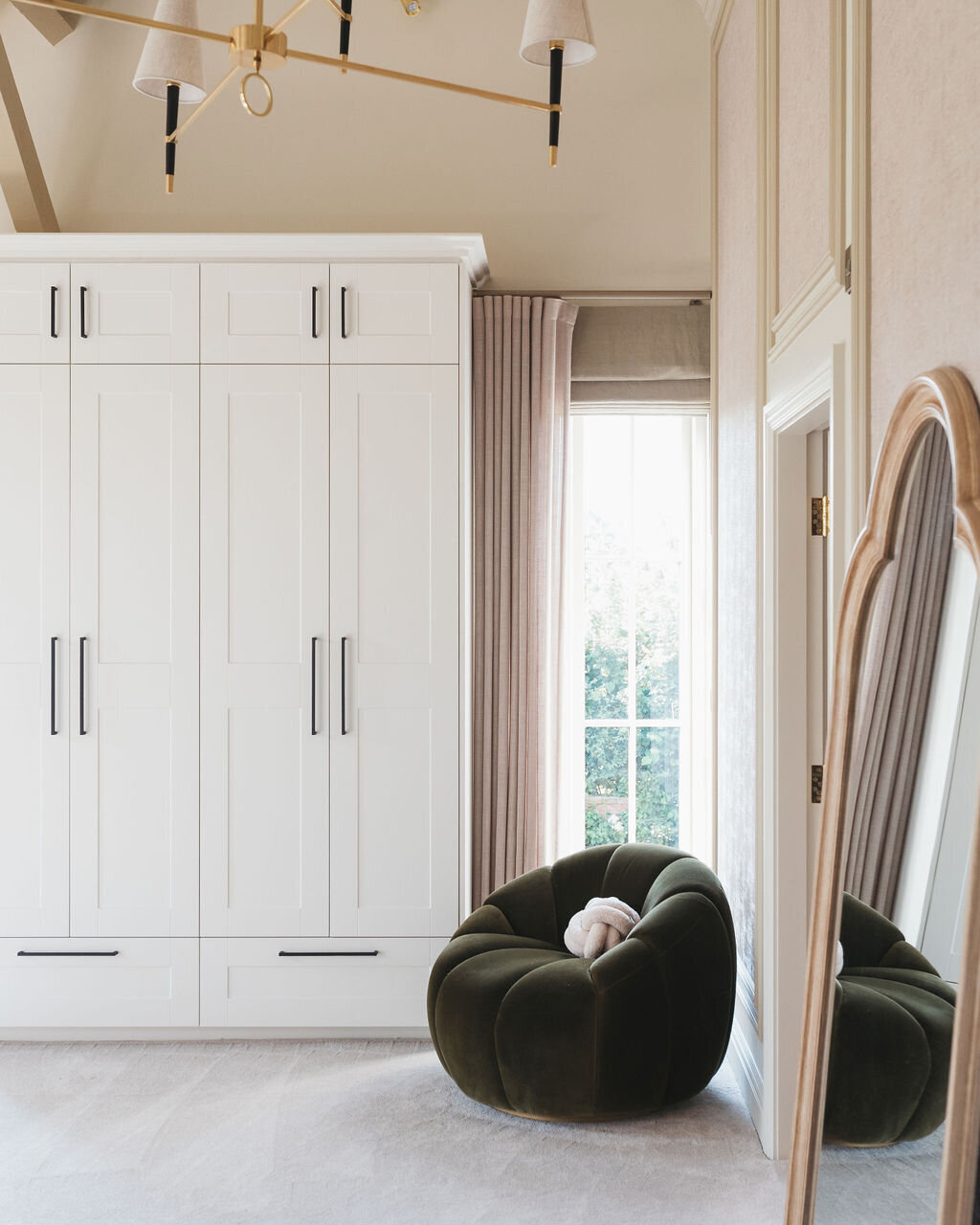 A cozy room features a corner with a plush, dark green chair holding a white pillow. Large white cabinetry with sleek black handles lines the wall. A tall arched gold mirror leans against the wall, reflecting natural light streaming through a window dressed with light curtains.