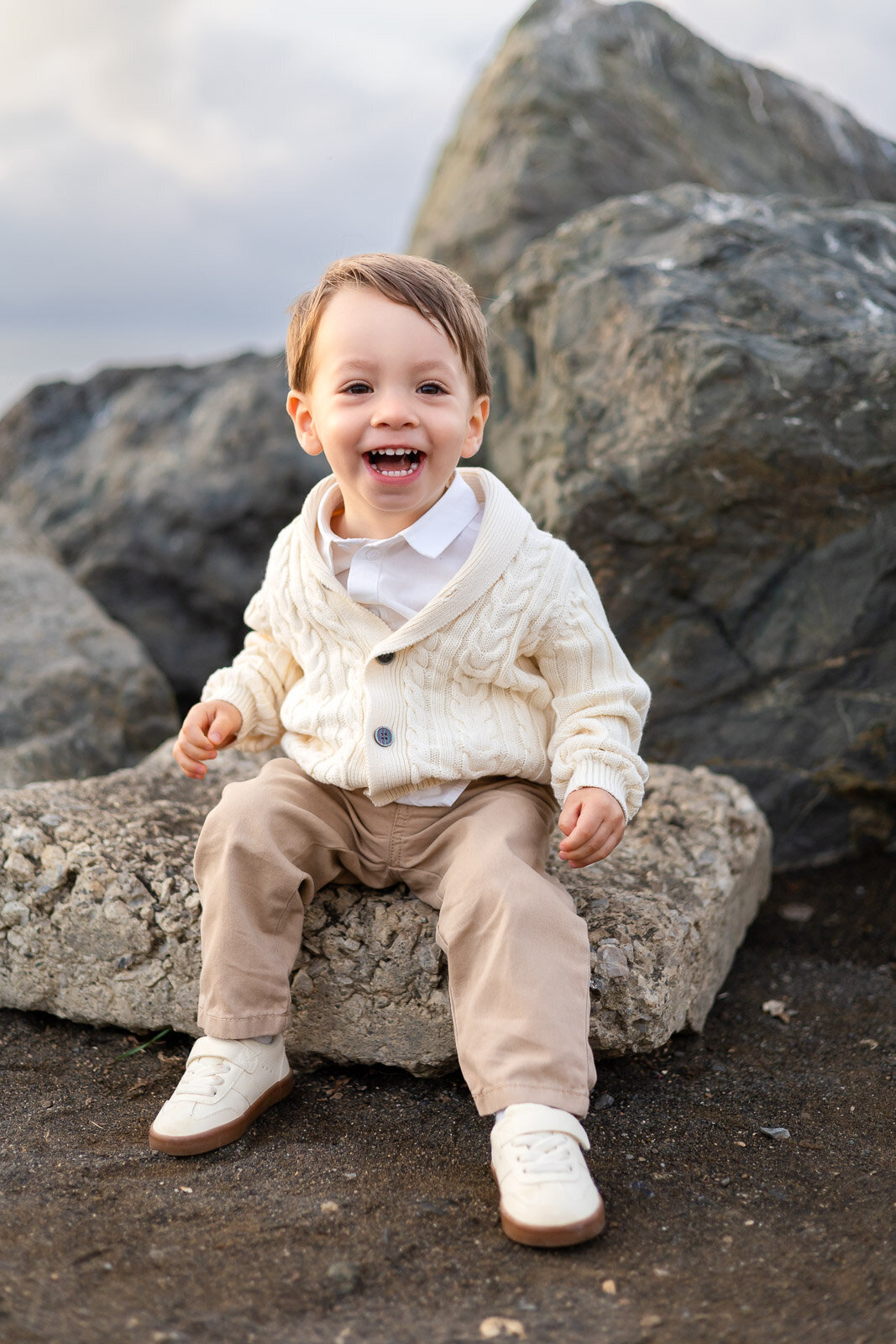 Young boy smiling in cream sweater sitting on beach rocks during San Mateo candid school photography lifestyle session – Ellobelle Photography