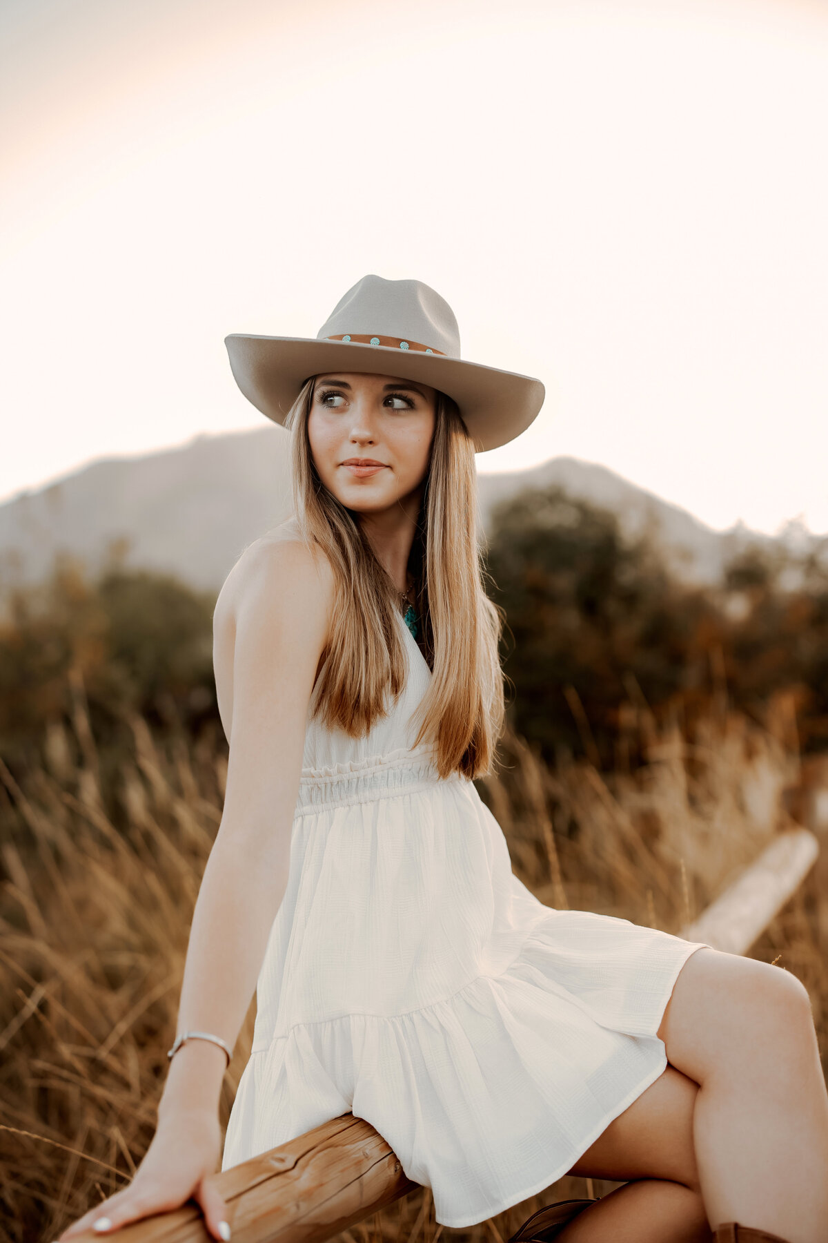 High school senior girls sits on wooden fence in a white dress and cowboy hat for her senior photos