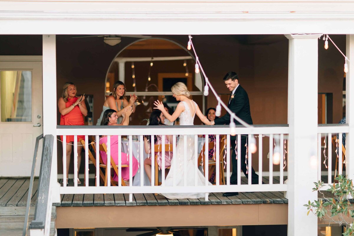 A bride and groom walk onto a balcony where guests seated at tables clap and cheer for them. The scene is festive, with string lights and the couple in wedding attire, creating a joyful celebration atmosphere.