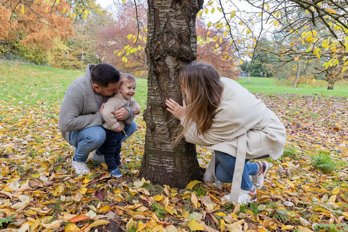 A happy family playing hide and seek around a tree in a park covered with autumn leaves.