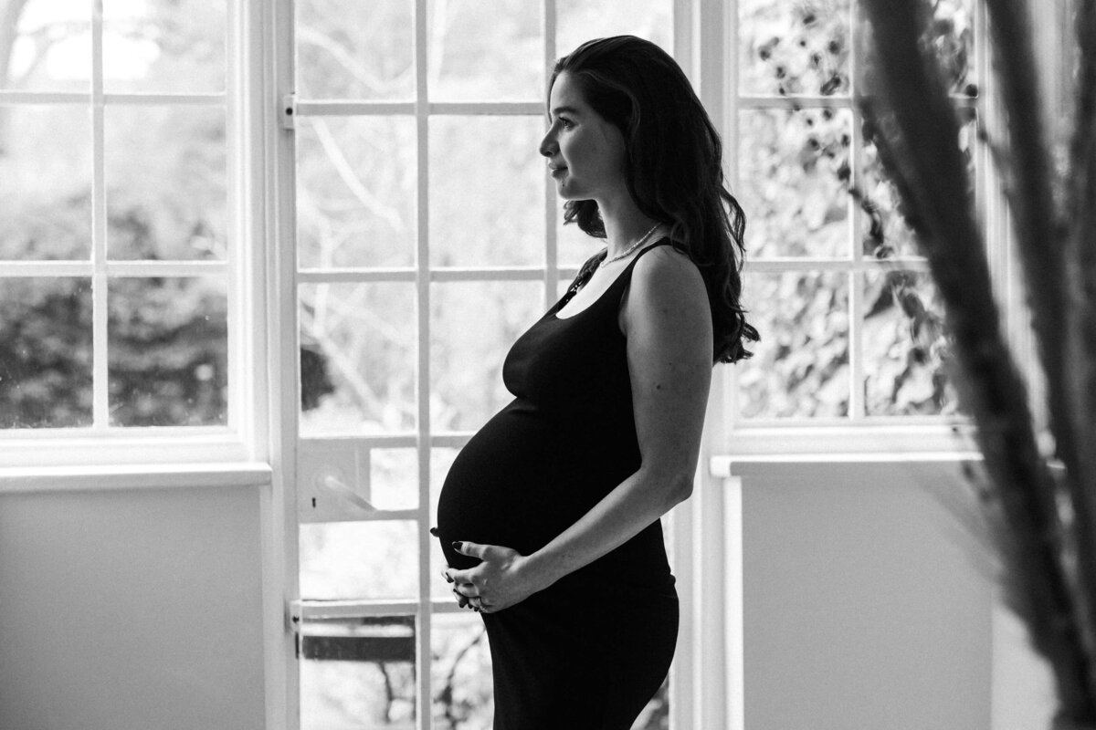 Pregnant woman wearing stretchy dress holds her baby bump in front of windows