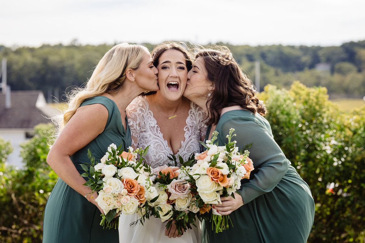 A bride in a lace gown laughing joyously as two bridesmaids in teal dresses kiss her cheeks, all holding bouquets with orange and white flowers.