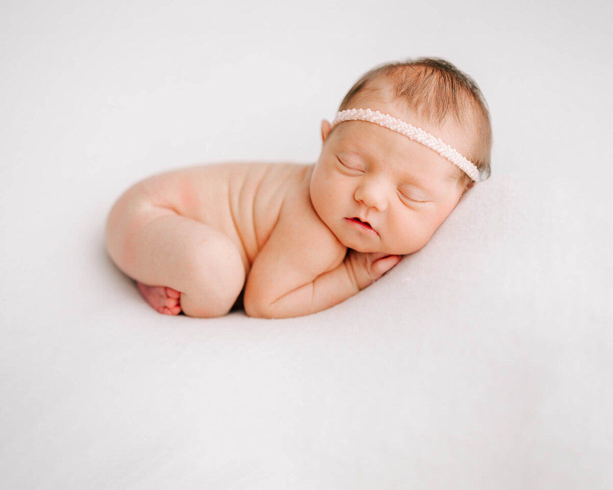 newborn infant girl sleeping on white blanket with her arms and legs under her and pink headband on