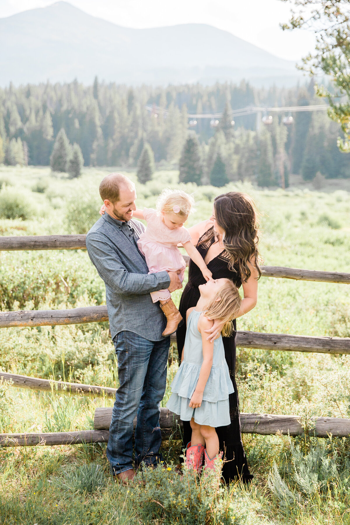 This family of four is all looking at one another and smiling. There is a log fence behind them with a wide open field and Breckenridge Mountain behind them