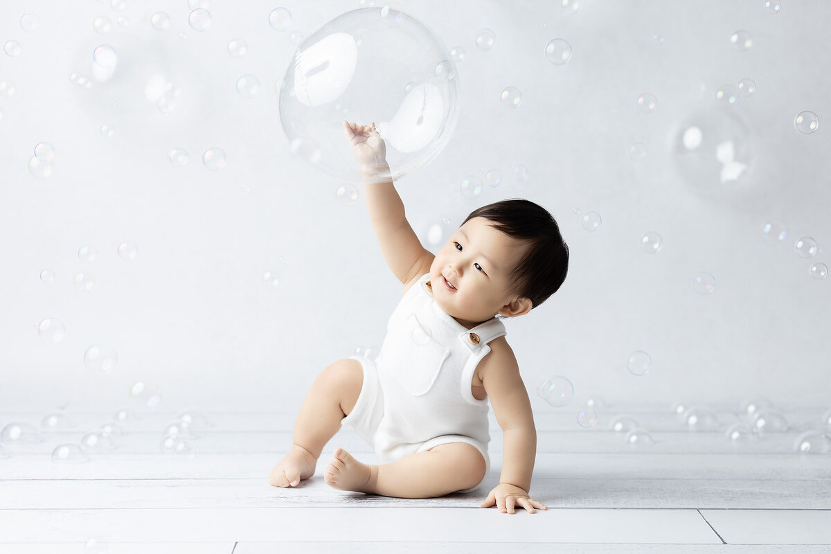 Dallas milestone photographer captures a baby in a white romper reaching out to touch a large bubble with a joyful smile, surrounded by floating bubbles, sitting on a light wooden floor.