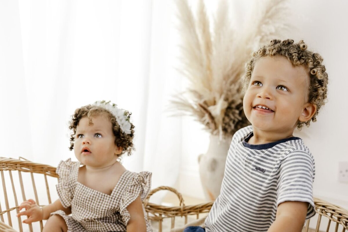 Two adorable siblings with curly hair, a younger one wearing a floral headband and a gingham outfit, and an older one in a striped shirt, sit in a wicker basket