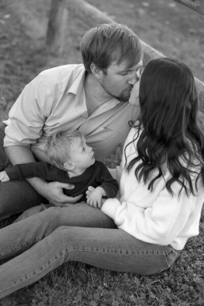 In a classic black and white portrait, a man and woman share a tender kiss while sitting on the grass. Their toddler, perched on the man's lap, gazes up at the woman. A wooden fence frames this intimate moment, captured with the artistry of New England's portrait photographers.
