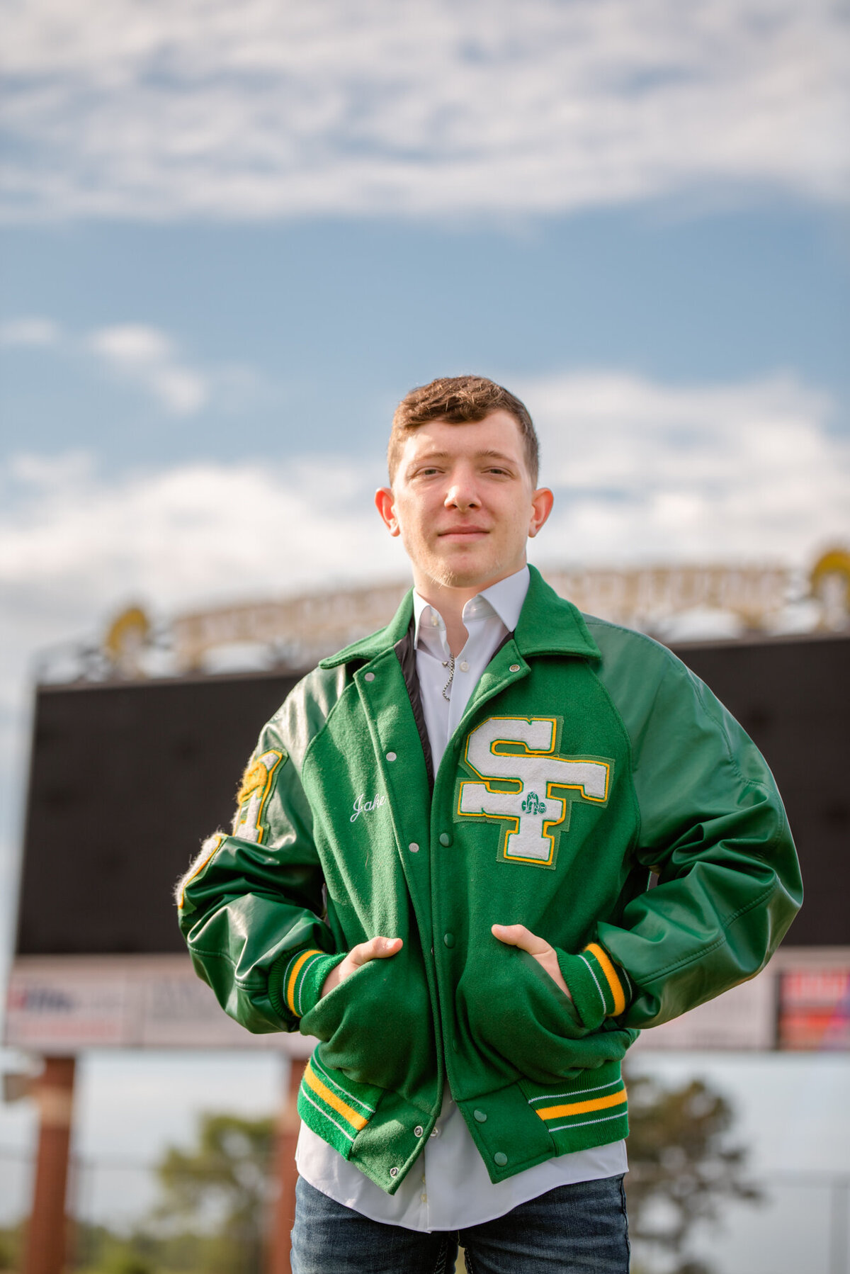 A senior wearing a green letterman stands in front of the football score board with a blue sky overhead.