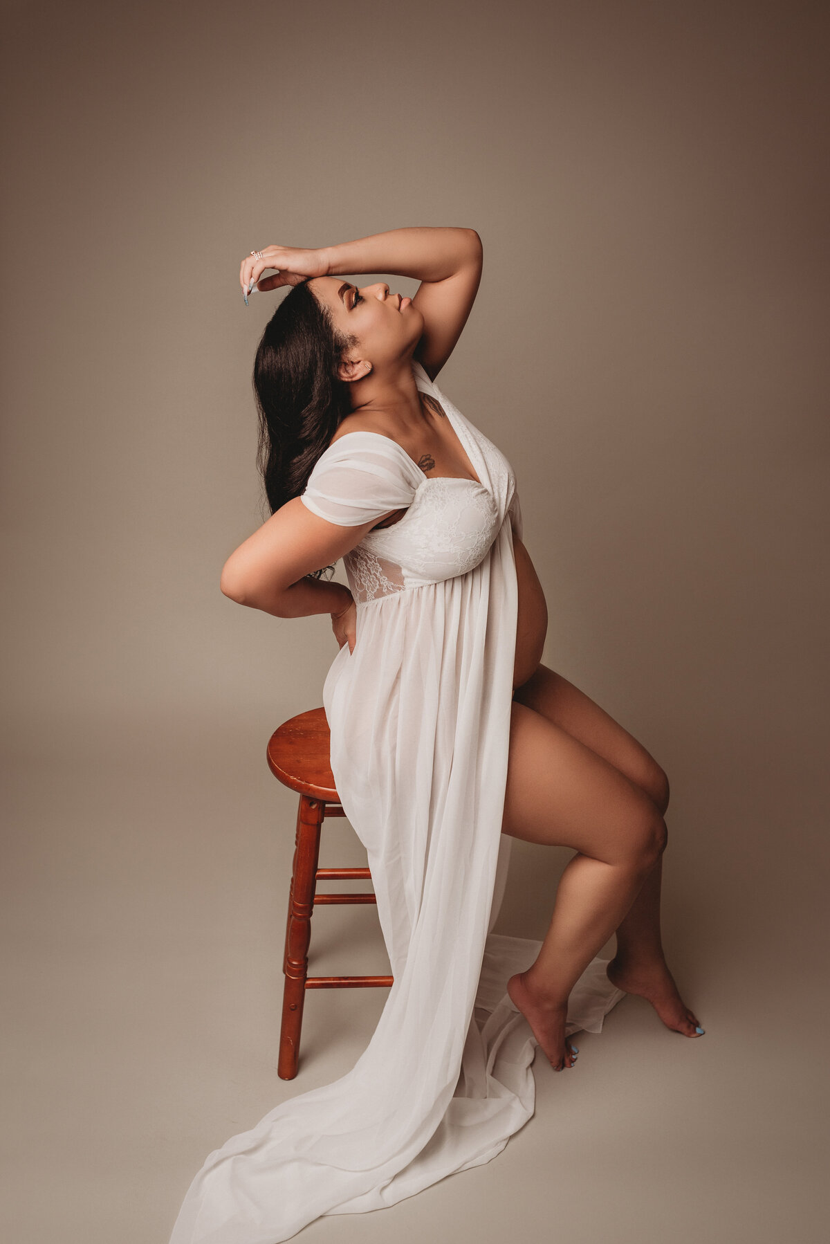 Pregnant woman in open white sheer dress exposing her belly sitting on a stool with hand on head