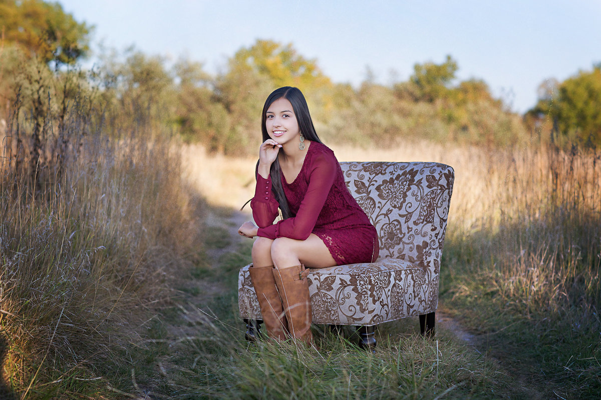 Senior girl in a burgundy dress sits on a chair in a meadow