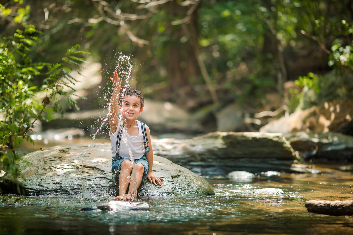 A young boy smiles and plays in a stream. He splashes water while sitting on a rock.