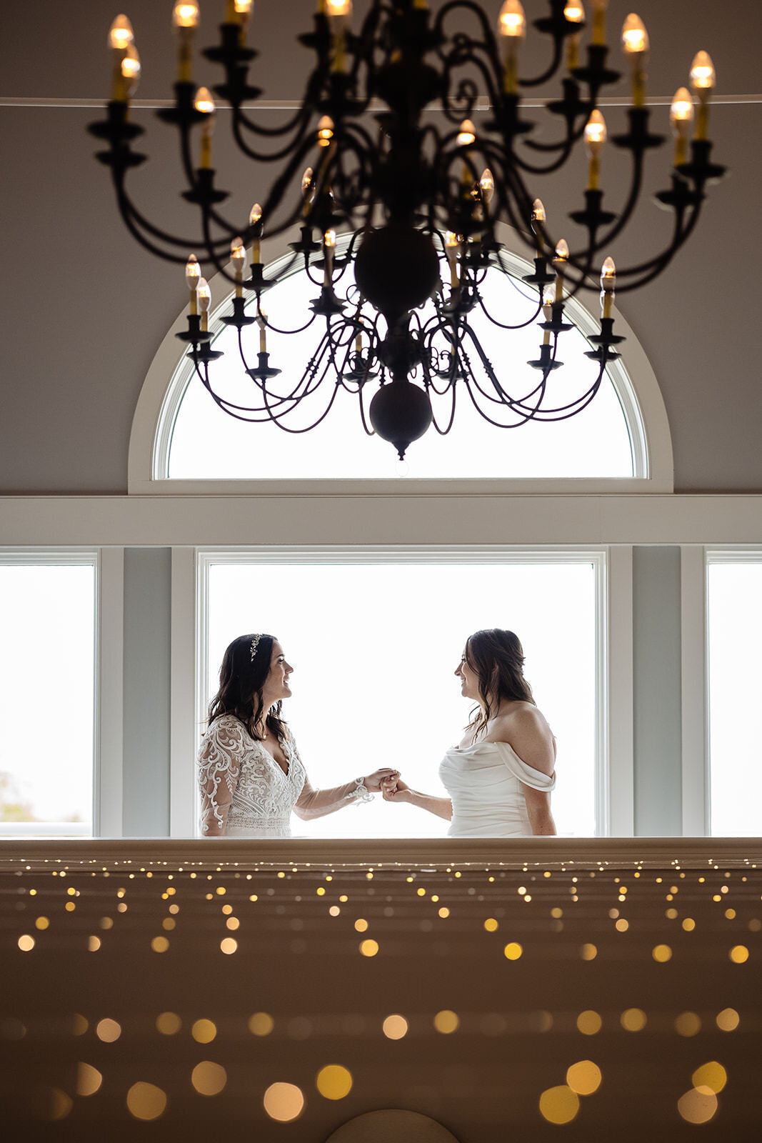 Two brides holding hands under a grand chandelier, silhouetted against a large window with soft golden light spilling in.
