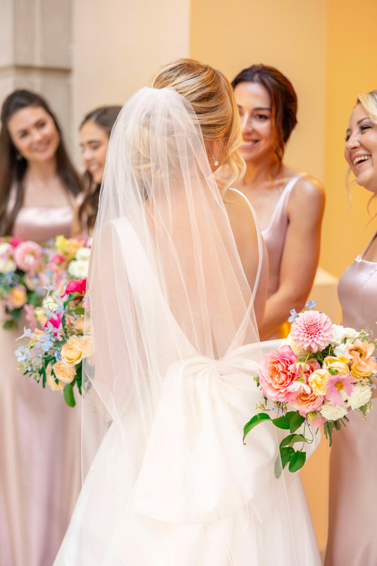 A bride in a white dress and veil is standing with her bridesmaids, who are holding colorful bouquets. The bridesmaids are wearing pale pink dresses and smiling. The setting is bright, with a warm, light background.