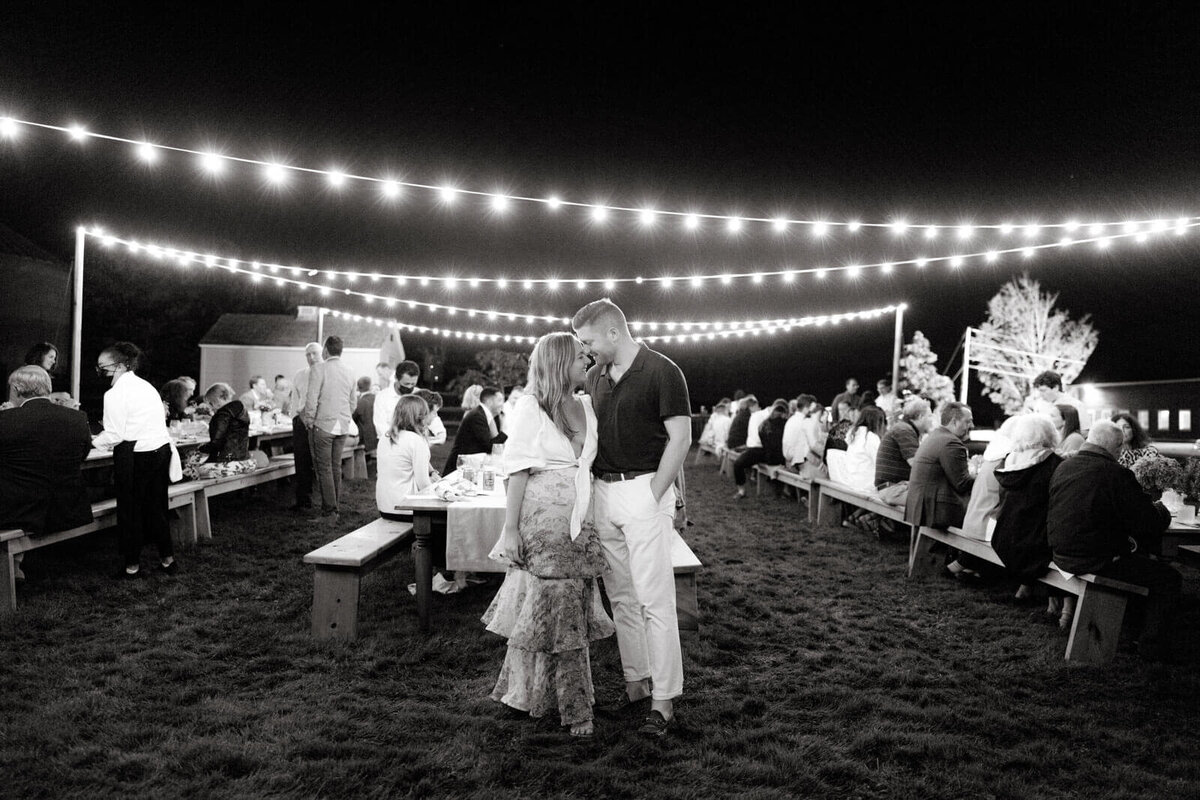 The bride and groom-to-be are standing close, heads touching, at a dinner outdoors, adorned with fairy lights, at Lion Rock Farms, CT.