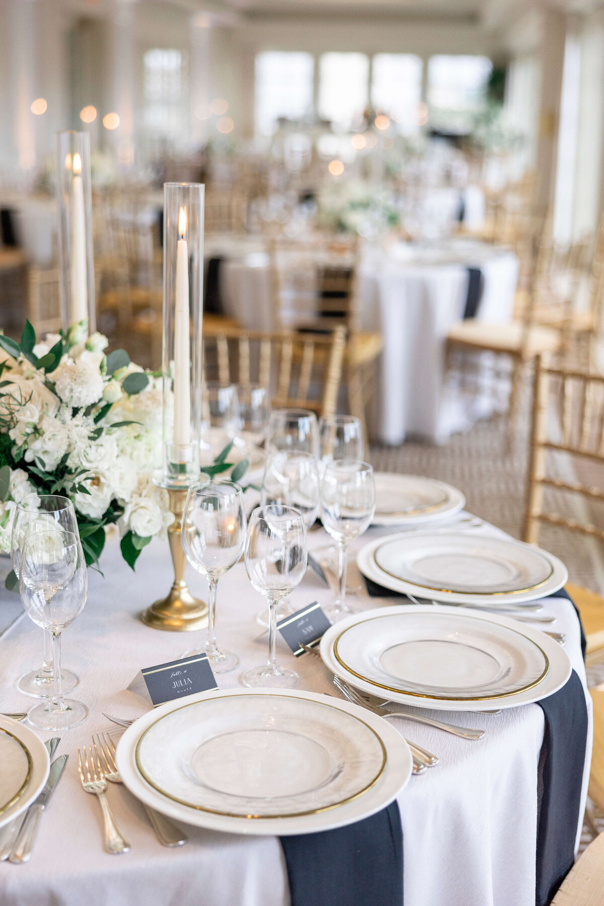 Elegant table setting with white plates, silver cutlery, and glasses on a white tablecloth with a black runner. Tall candles and a floral centerpiece with white flowers and greenery are in the middle. Gold chairs and more tables in the background.