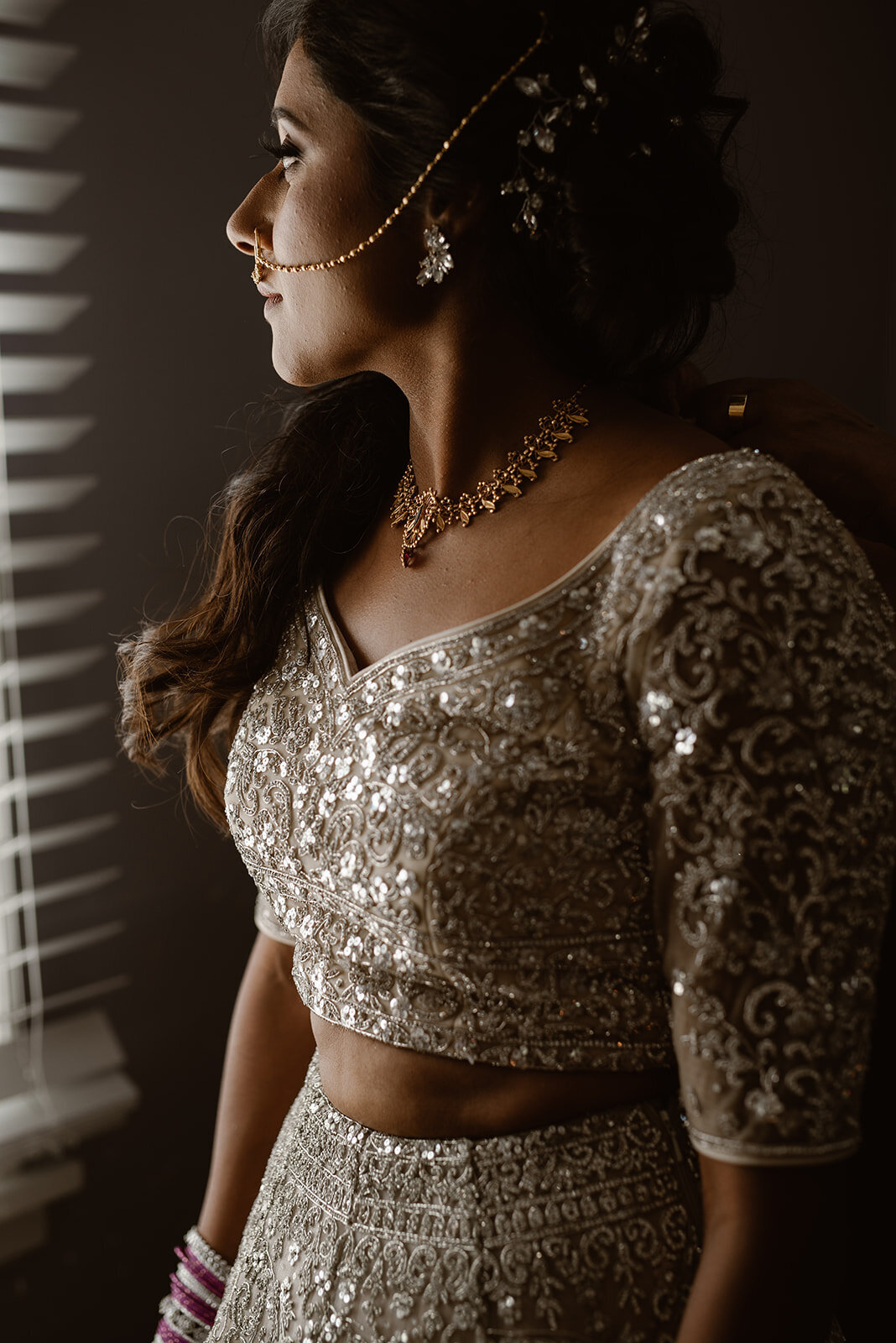 A bride in an intricately embroidered lehenga stands near a window, her silhouette softly illuminated.