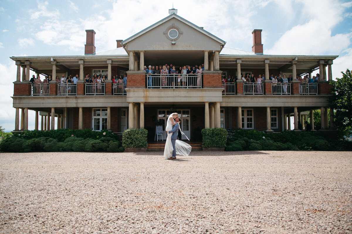 A Bride and groom embrace in front of a big manor house , all their guests are on the balcony of the house