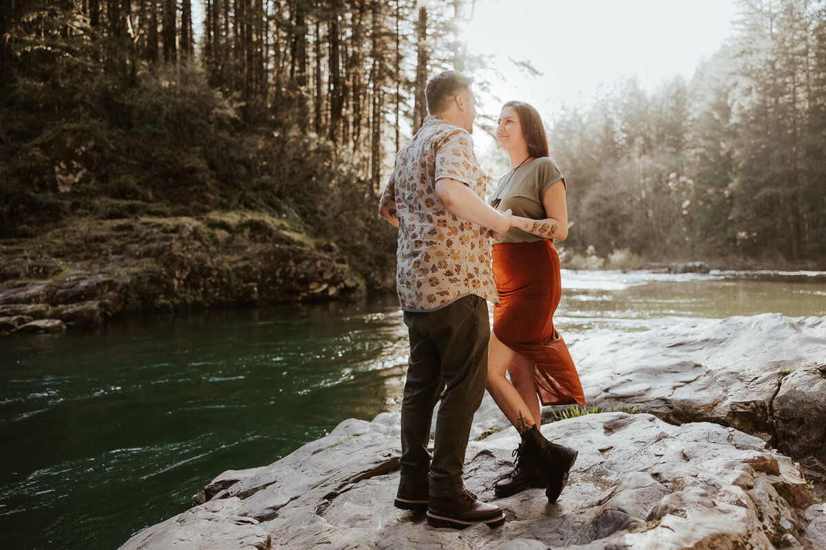 A happy couple stands on a rock by the river holding hands at sunset