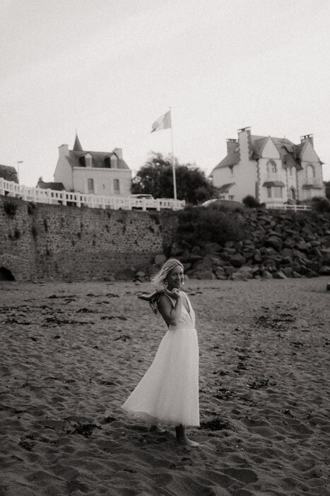 Mariée posant chaussures sur son épaule à la plage. Photographie en noir et blanc par kamescop, photographe vidéaste mariage à Saint Quay Portrieux, Côtes d'Armor, Bretagne.