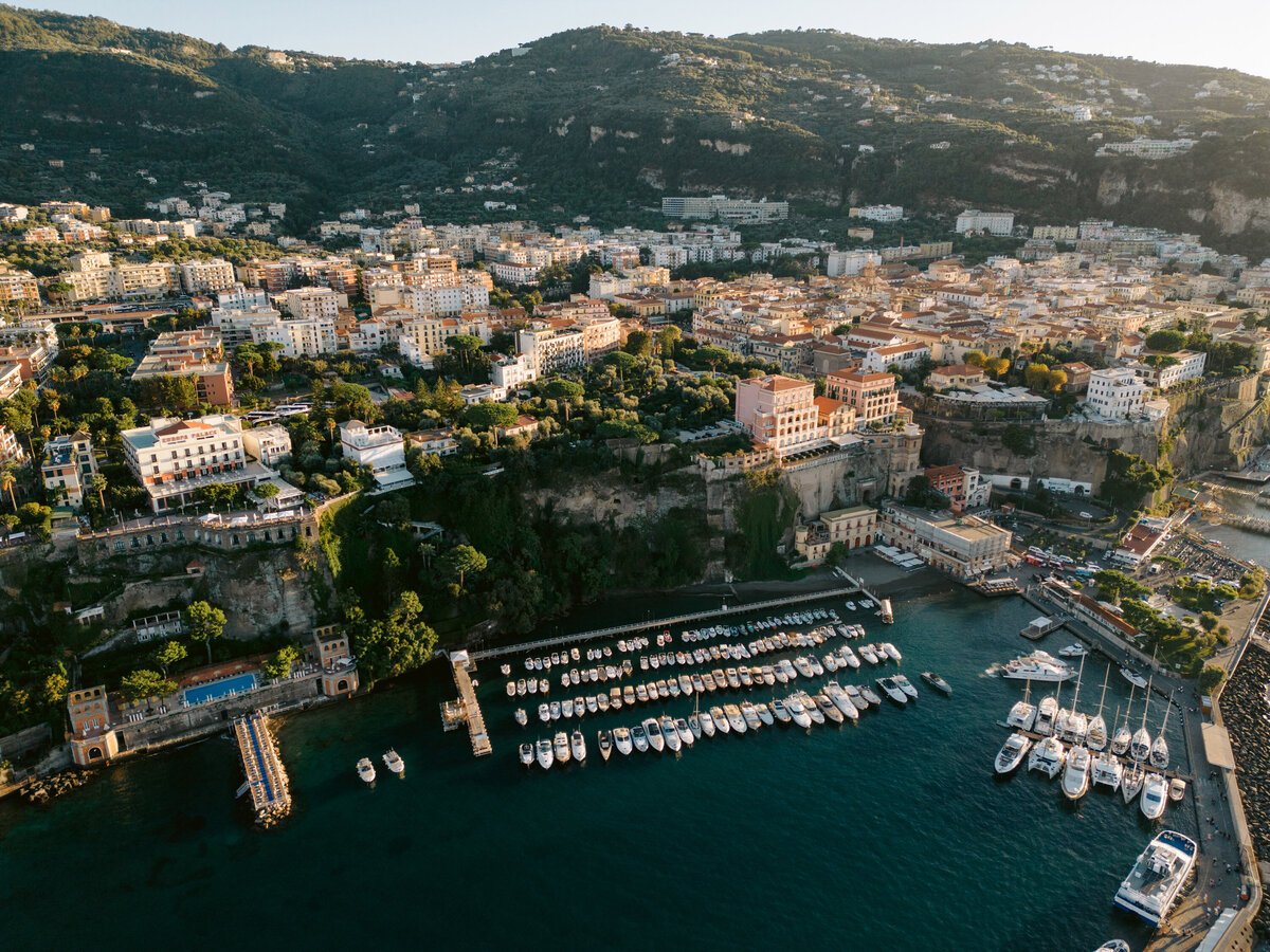 Aerial view of Sorrento’s coastline, showcasing a picturesque harbor filled with boats, historic buildings perched on cliffs, and lush hills in the background, captured during a destination wedding.