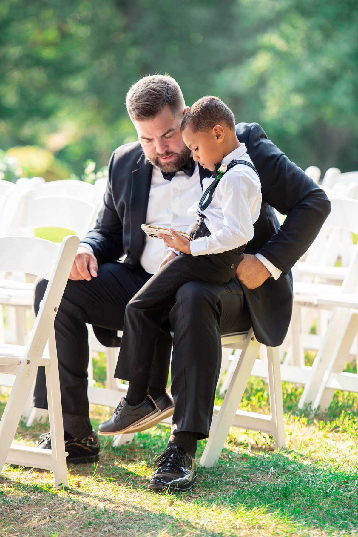 A man in a suit sits on a white chair outdoors, holding a young boy dressed in formal attire with suspenders. The boy is focused on a smartphone. The scene is set in a grassy area with white chairs in the background.