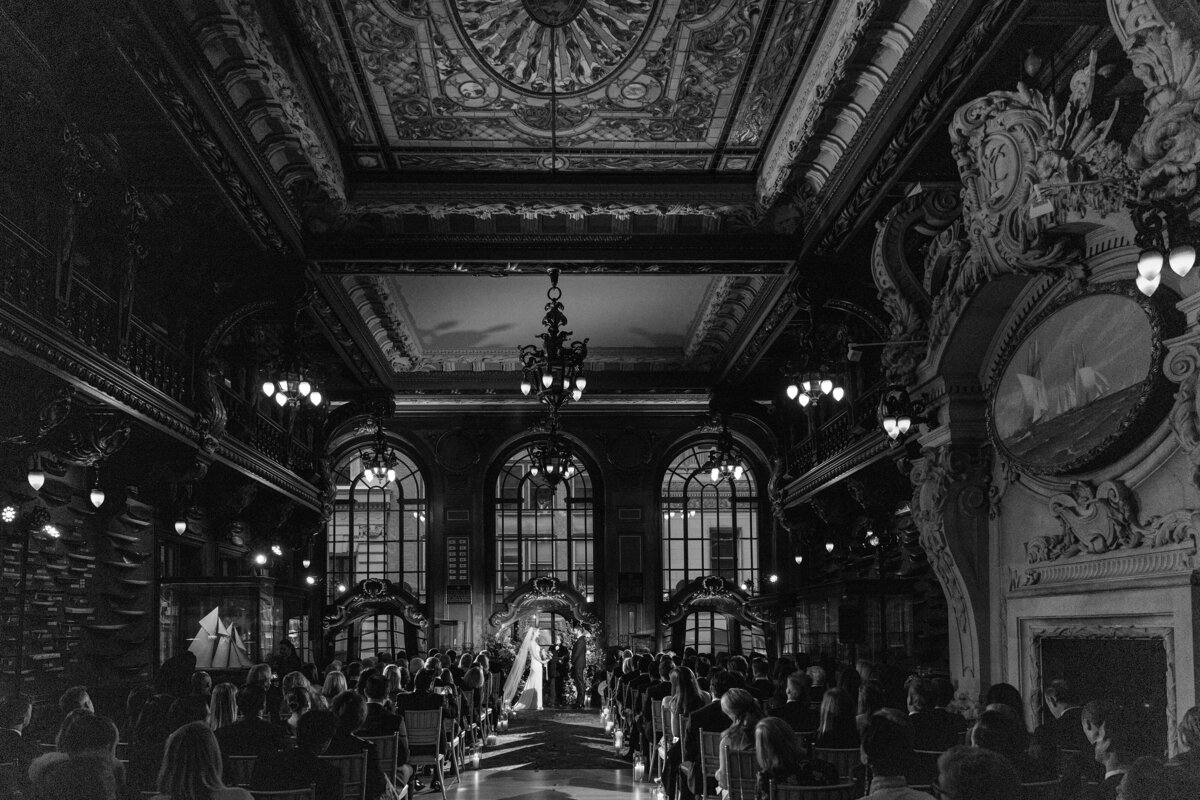 Black and white photograph of a wedding ceremony inside the New York Yacht Club’s ornate Model Room, with vaulted stained glass ceilings, intricate woodwork, and a crowd of guests seated facing the couple standing at the altar.