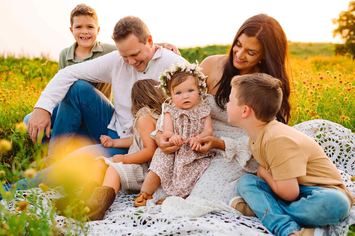 The skilled family photographer in Albuquerque captures a joyful family moment on a mountain. The large family is sitting on a white blanket, with the parents engaging and playing with their kids.