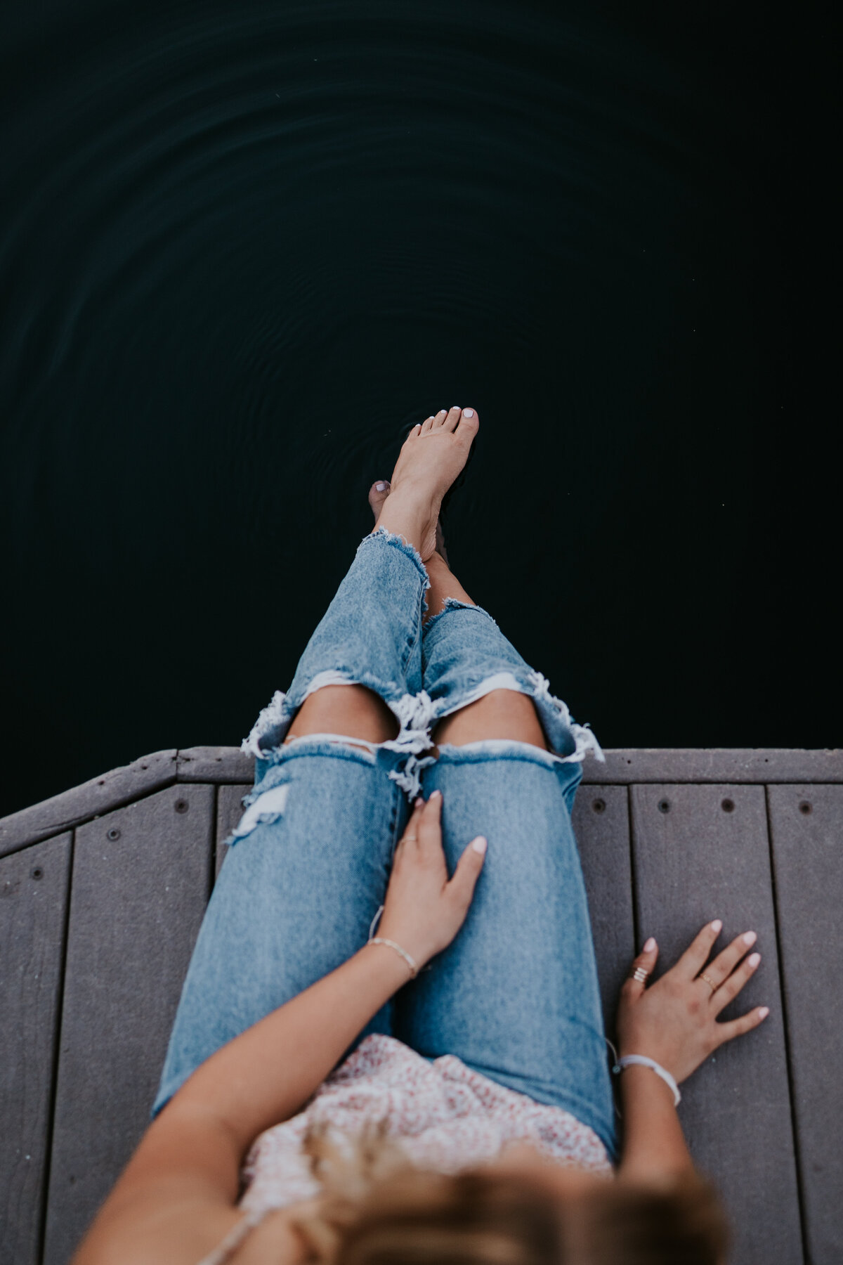 Camera looks down on young woman while she sits on dock dipping her toes in water.