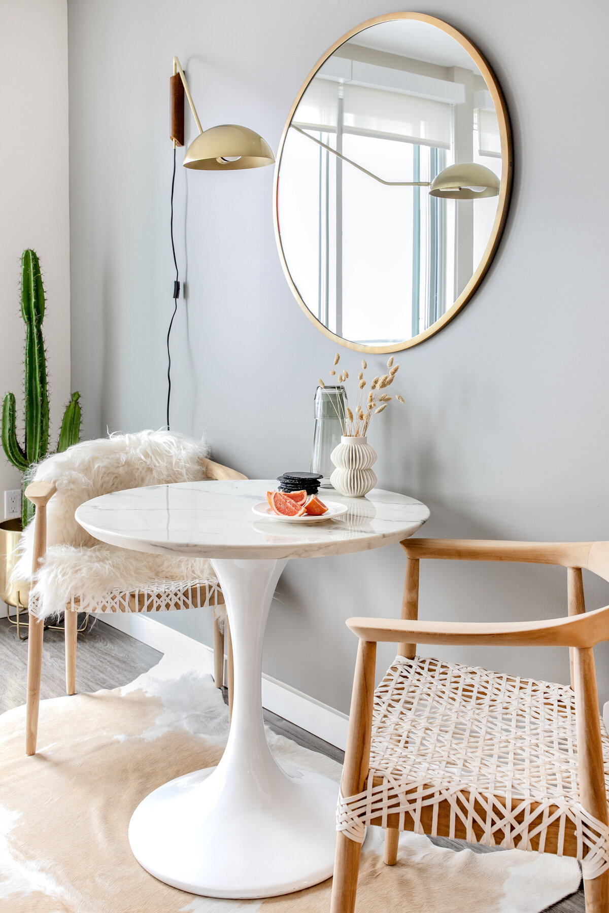 White marble Saarinen tulip dining table alongside wooden dining chairs with white leather woven seats. Round brass mirror and  brass sconce light hanging above