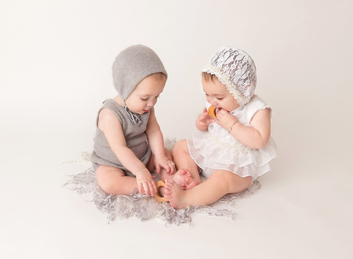 Twin brother and sister are sitting for a 6-month milestone photoshoot in Brooklyn, NY photo studio. Brother is wearing a taupe knit romper and bonnet and sister is wearing a white lace romper and bonnet. They are playing with wooden toys.