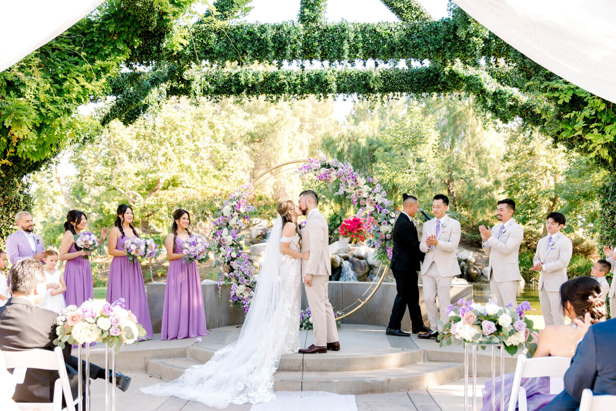 A couple shares a first kiss at an outdoor wedding under a floral arch, surrounded by bridesmaids in lavender and groomsmen in light suits, amid lush greenery.