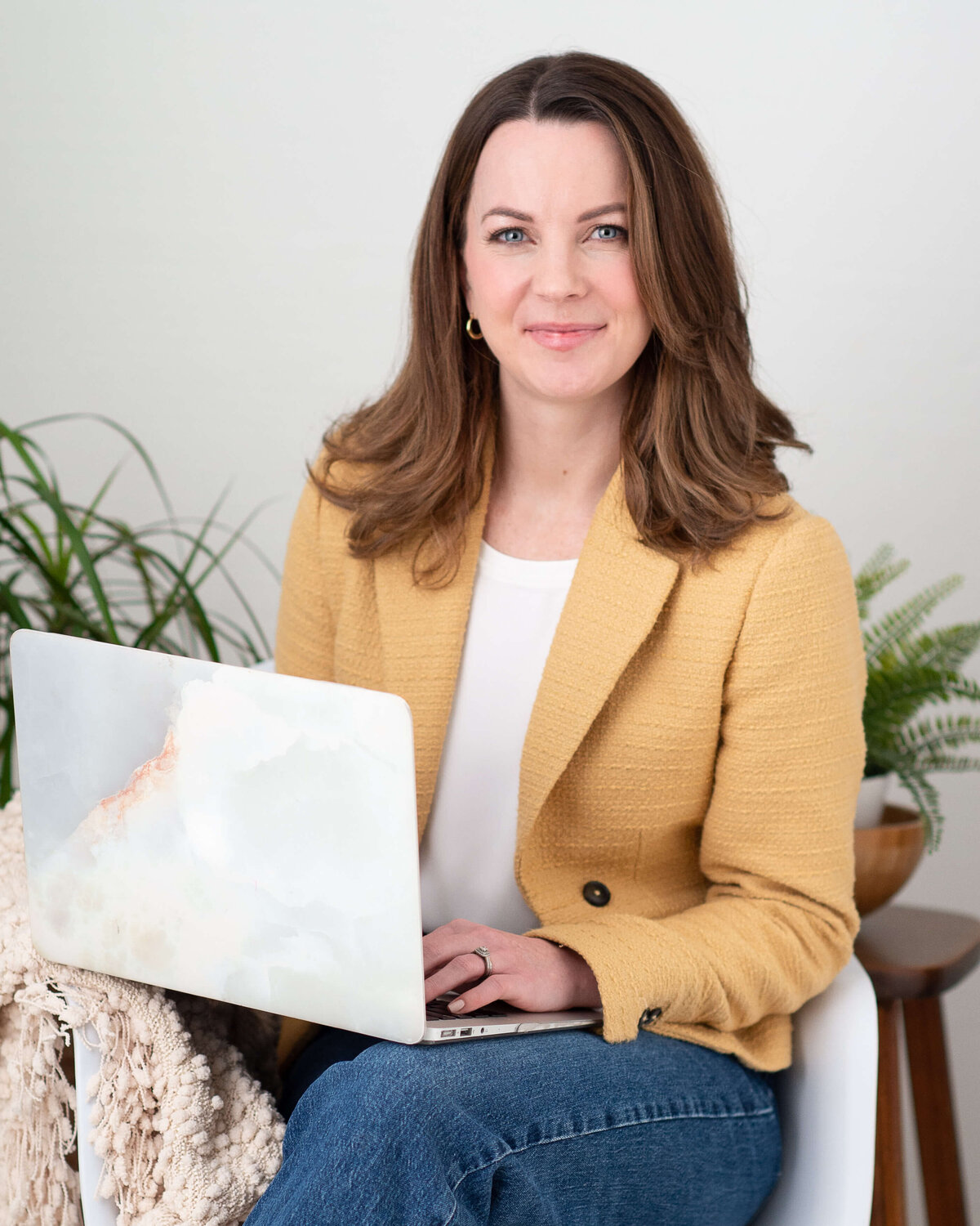 a woman in a yellow jacket sitting in a white chair typing at her laptop.  Captured by Ottawa branding photographer JEMMAN Photography