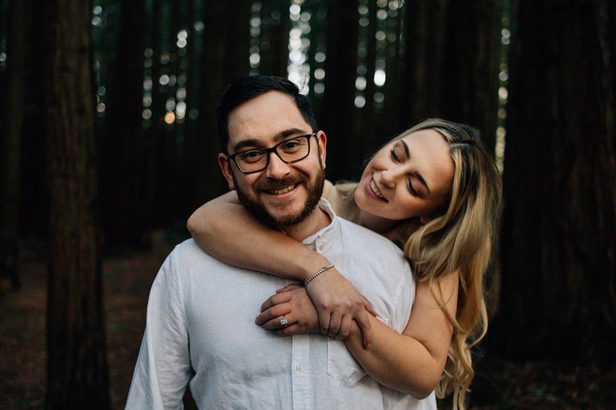Woman holding fiancé from behind, during engagement photoshoot in Redwood Forest, by Delanie And So I Don't Forget Photography