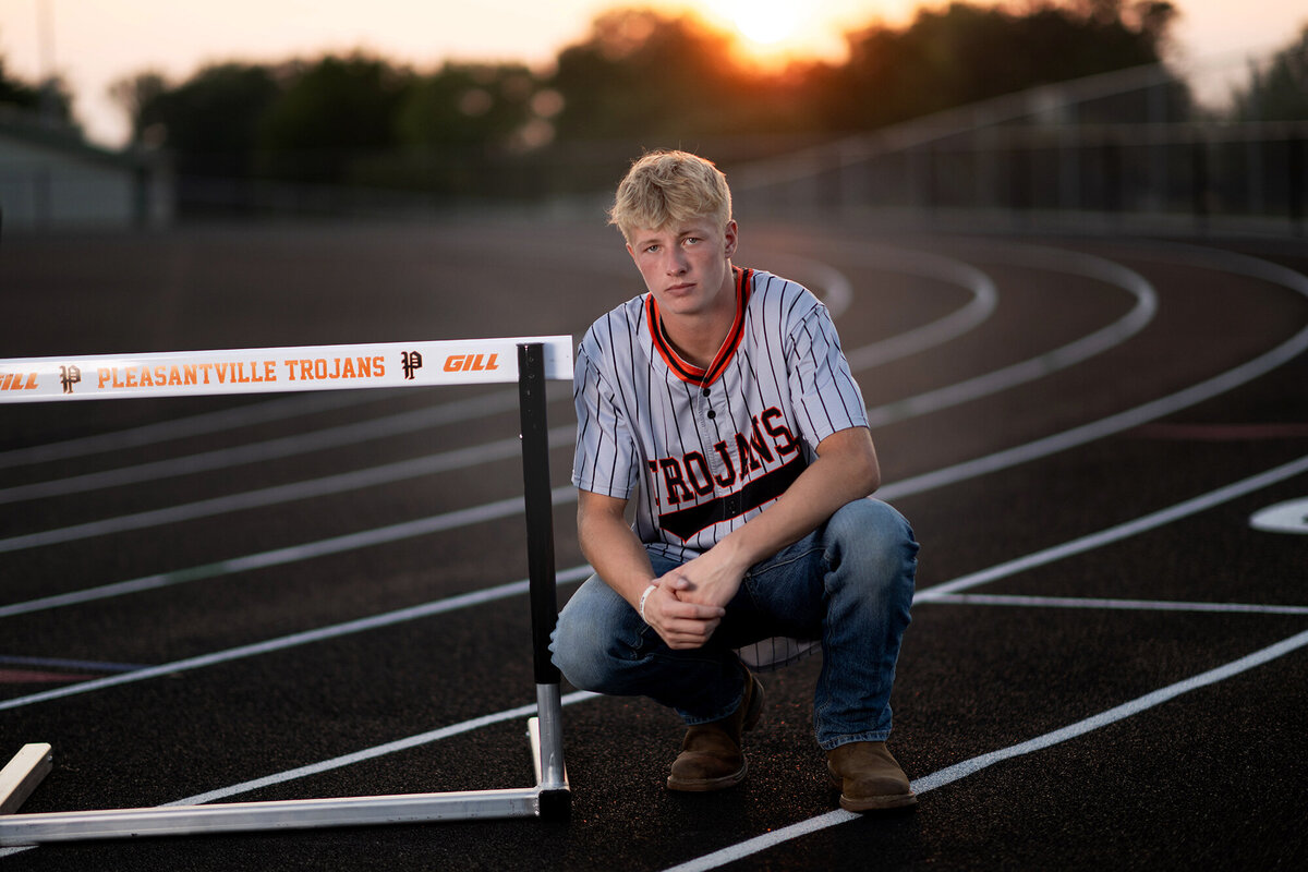 A high school senior squats on a track at sunset with a hurdle for his Des Moines Senior Photographer