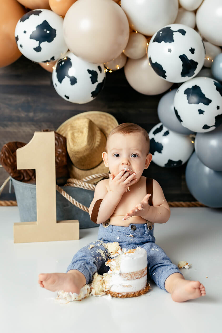 A baby sits on the floor, wearing jeans and suspenders, eating a small cake with their hands. A large number "1" and cow-patterned balloons surround them, creating a festive atmosphere. A straw hat and rope are in the background.